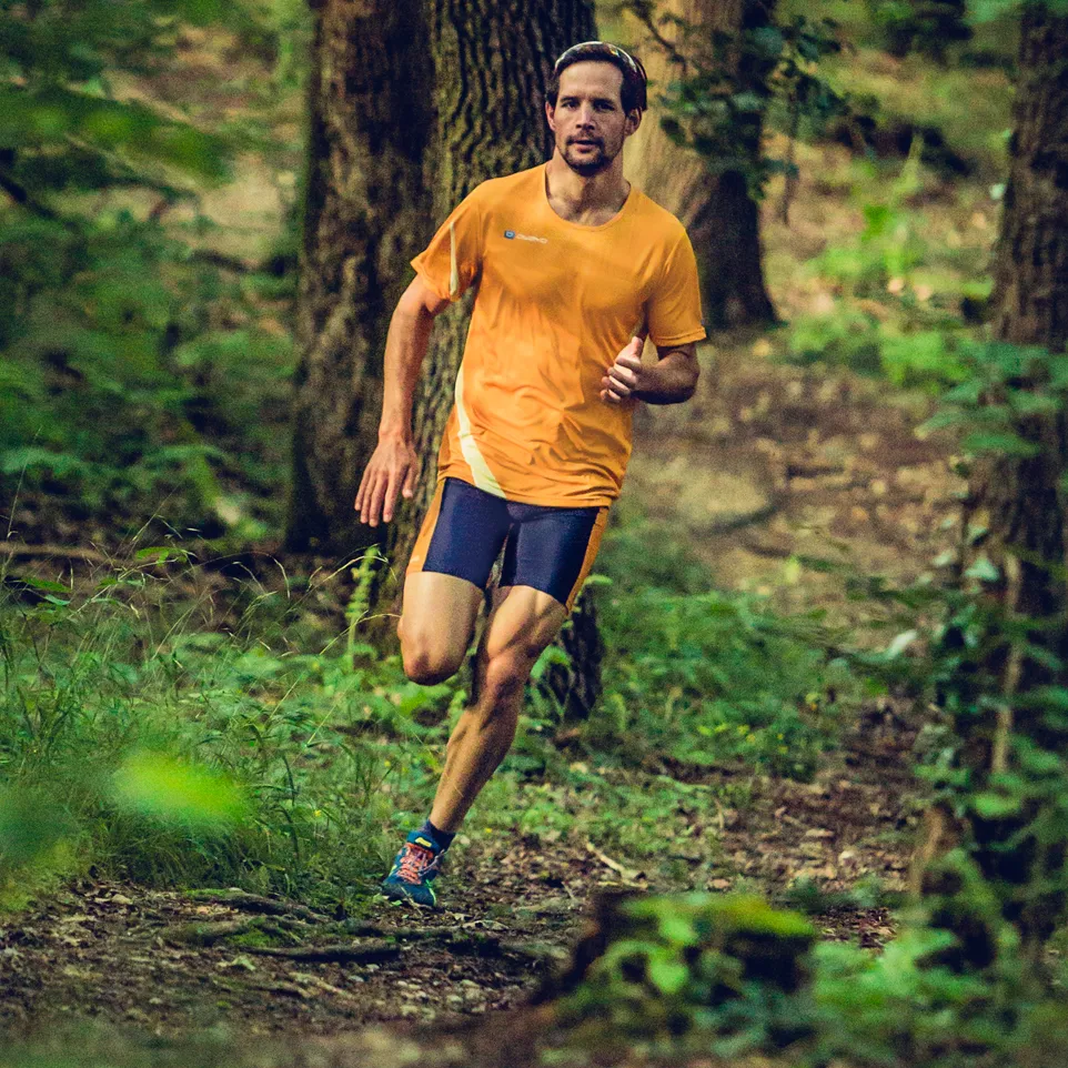 Ein Mann läuft auf einem Waldweg in einem orangefarbenen Lauftrikot und dunkelblauen Laufshorts.